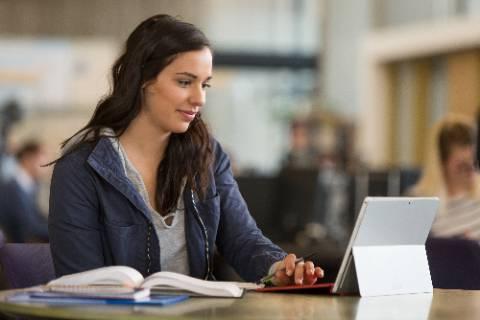 A female presenting person sitting in a library with books and an open laptop.