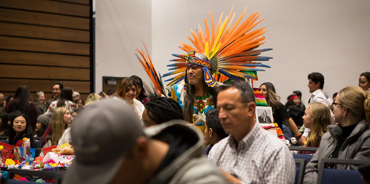 Photo of people sitting at tables and a man wearing a penacho, an aztec feathered headdress